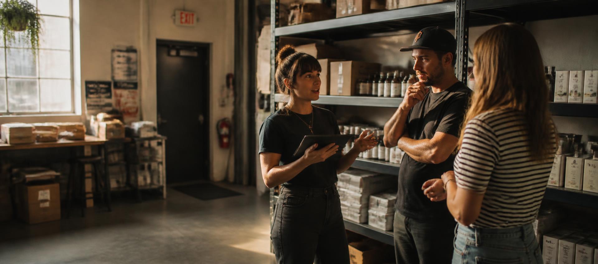 A small team on a shop floor mid-conversation, one holding a tablet, stock behind them