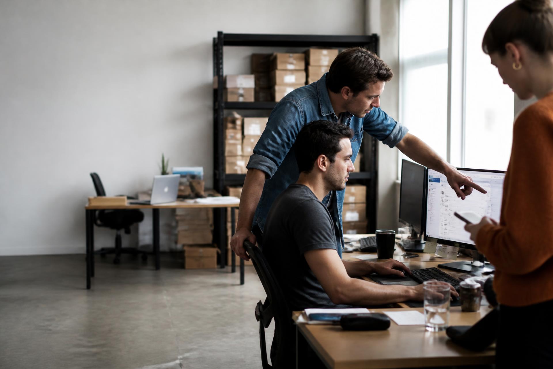 Three ecommerce colleagues gathered around one laptop in a warm sunlit workspace, all looking at the same screen